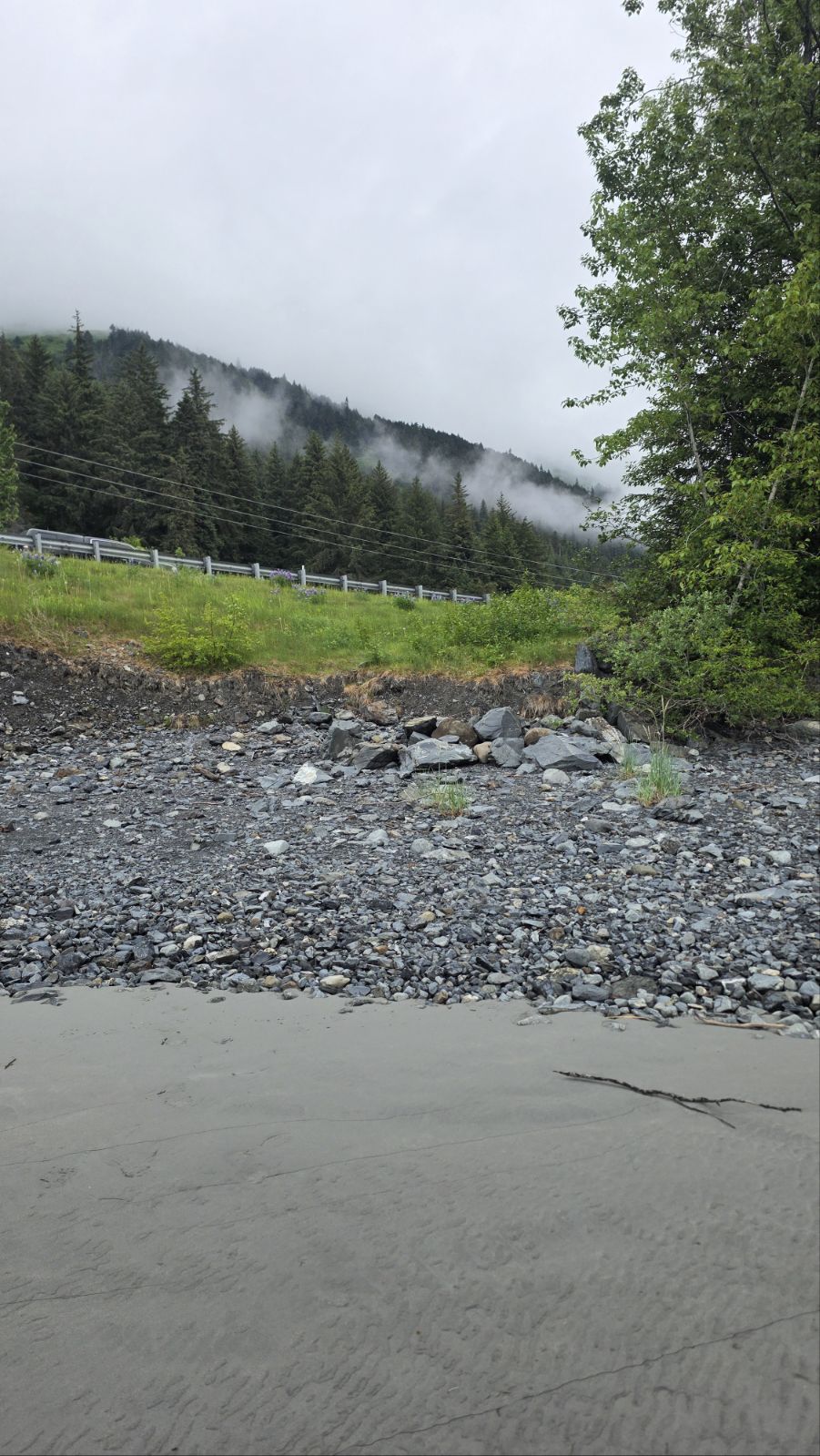 Riprap and loose rock between the Seward Highway guardrail and the tidal flats at Gunmount, with fog on the forested hillside above.