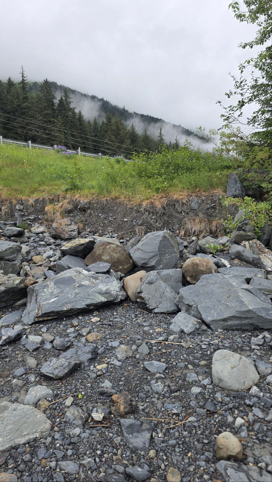 Close view of the broken rock armoring at the edge of the embankment, with the highway guardrail on the slope above.
