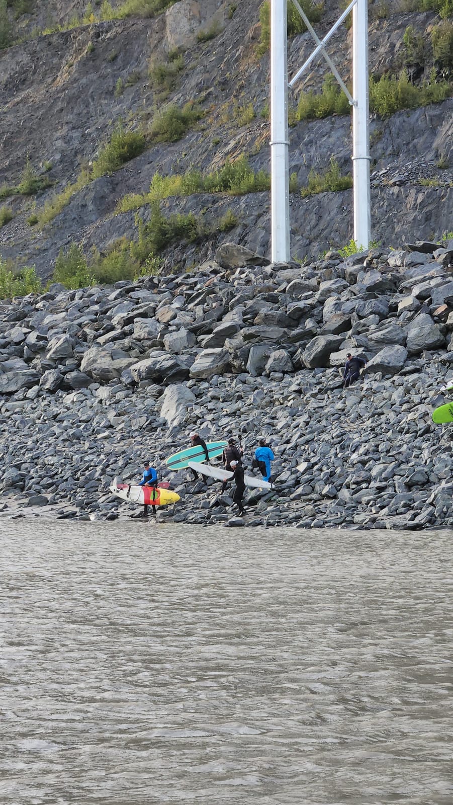 Bore-tide surfers carrying boards across the riprap shoreline of Turnagain Arm below the Seward Highway bridge near Girdwood, Alaska